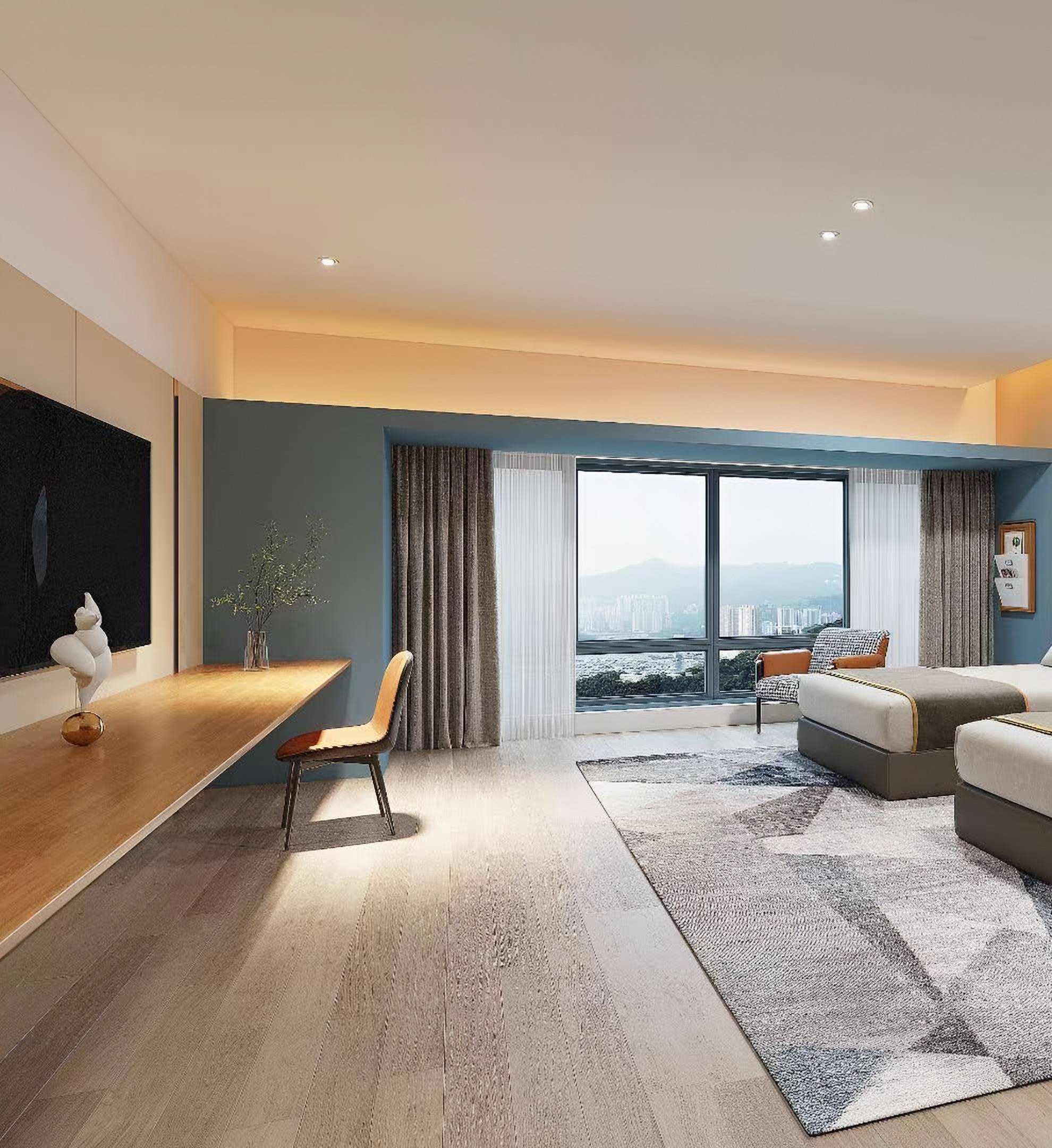 Hotel guest room with a long timber desk, a blue-framed window bay onto the city and hills, and an upholstered headboard